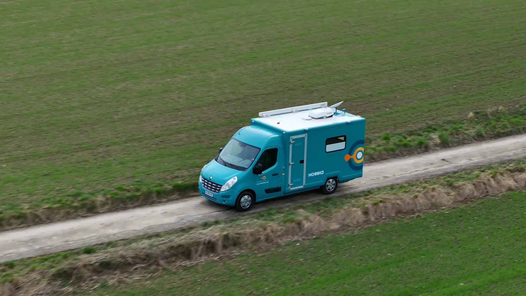 Seen from above, a turquoise Médicobus drives alone on a dirt road between two fields
